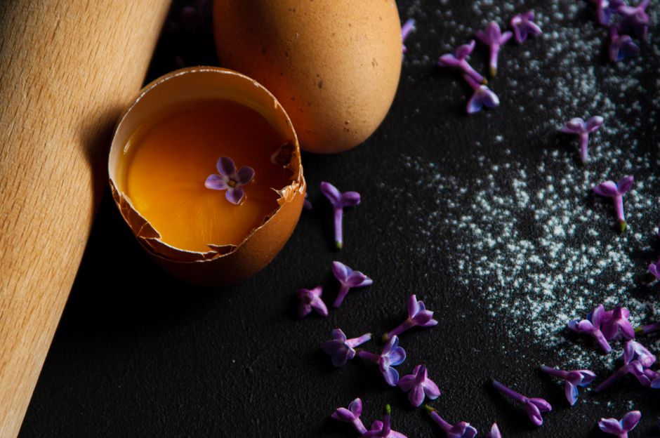 Fleurs comestibles et œuf cassé sur un plan de travail, avec un rouleau à pâtisserie et du sucre en poudre.