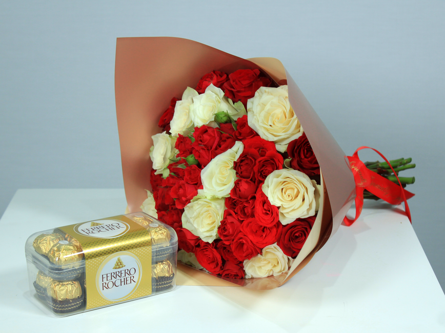 Bouquet de roses rouges et blanches accompagné d'une boîte de chocolats Ferrero Rocher sur une table.