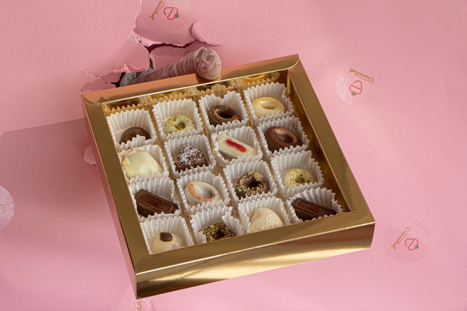 Coffret de chocolats assortis sur fond rose, contenant 16 pralines élégamment présentées.