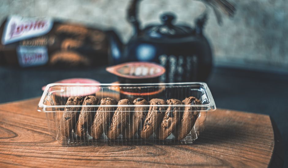 Biscuits gourmands en boîte transparente sur une planche en bois, avec des pots de crème en arrière-plan.