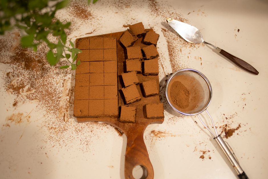 Brownies fondants découpés en carrés sur une planche en bois, avec de la poudre de cacao et un tamis à proximité.
