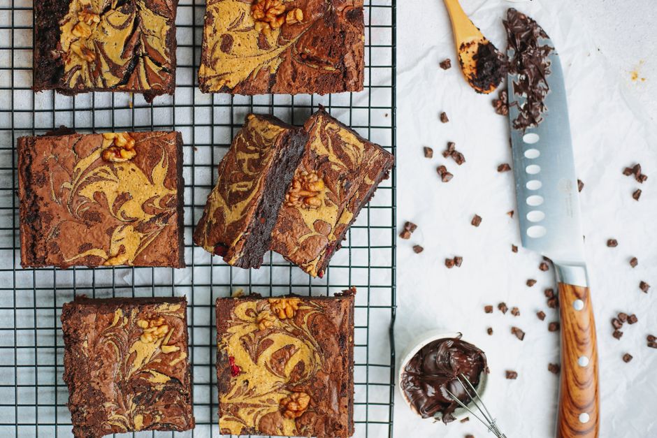 Brownies fondants découpés sur une grille, décorés de marbrures et de noix, avec des éclats de chocolat à côté.